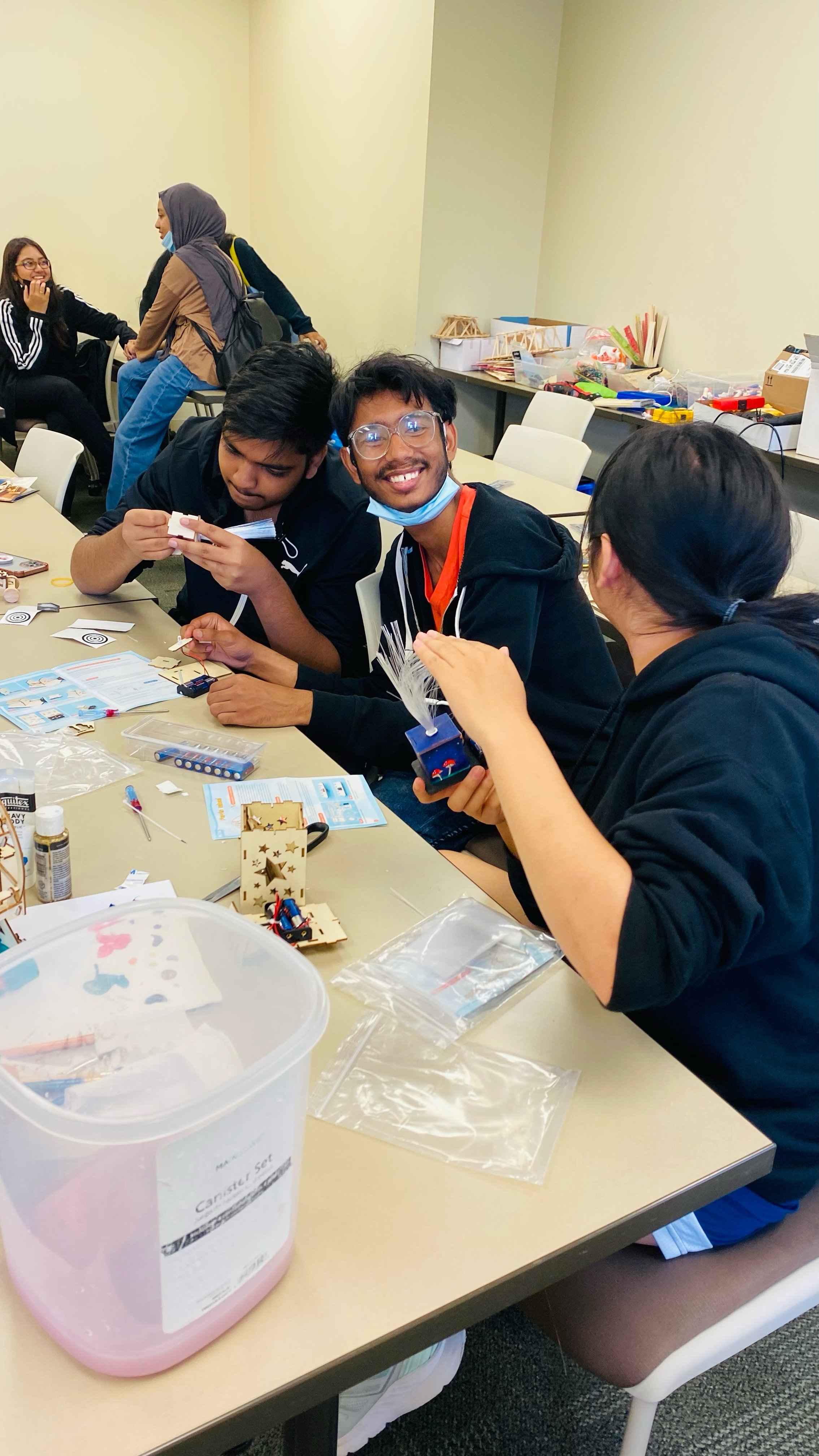 Group of young people working on a creative project at a table, surrounded by art supplies and tools, in a classroom or workshop setting.