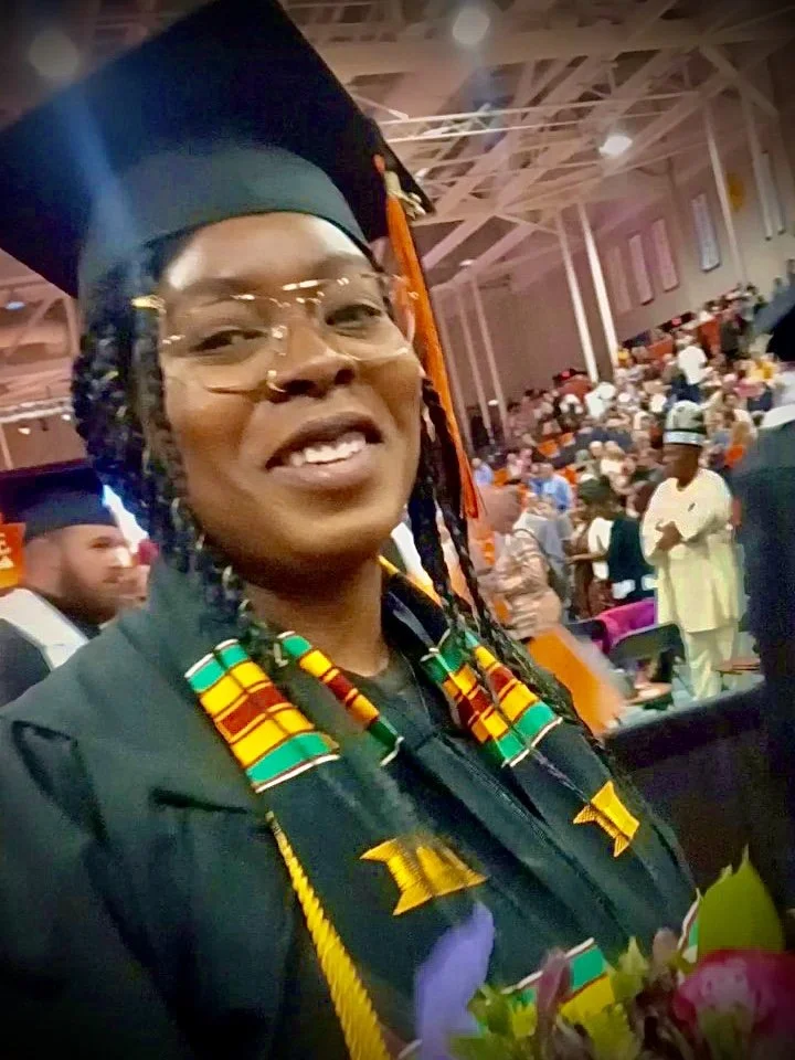 Woman in graduation cap and gown smiling at a ceremony, with many people in the background.