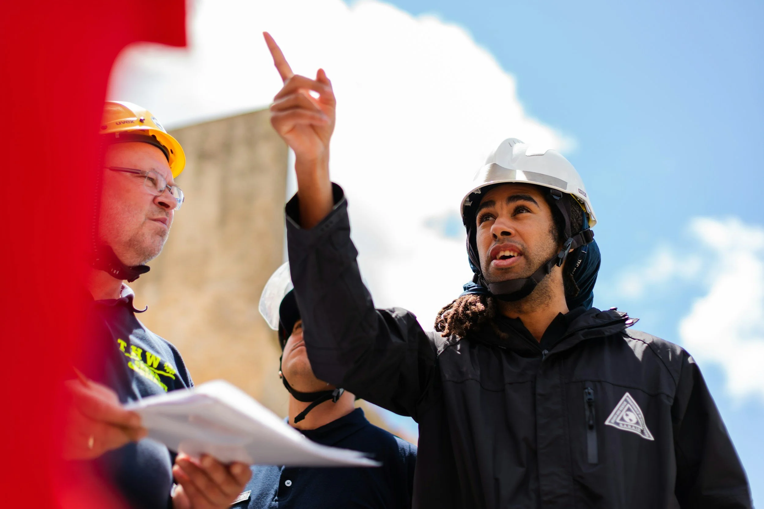 Two men wearing safety helmets and jackets are outdoors under a cloudy sky. One man is pointing or gesturing while the other is looking on and holding papers. A red object is partially visible in the foreground.