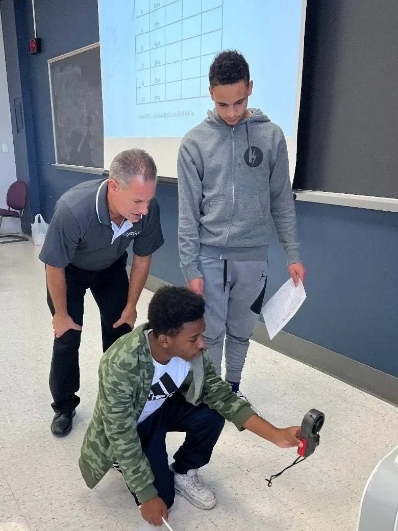 A teacher and two students in a classroom, one student is crouching and holding a handheld device, while the other student is standing and holding a paper. There is a large screen and a blackboard behind them.