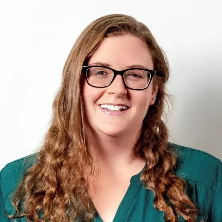 A woman with long, wavy red hair, wearing glasses and a green top, smiling against a plain white background.