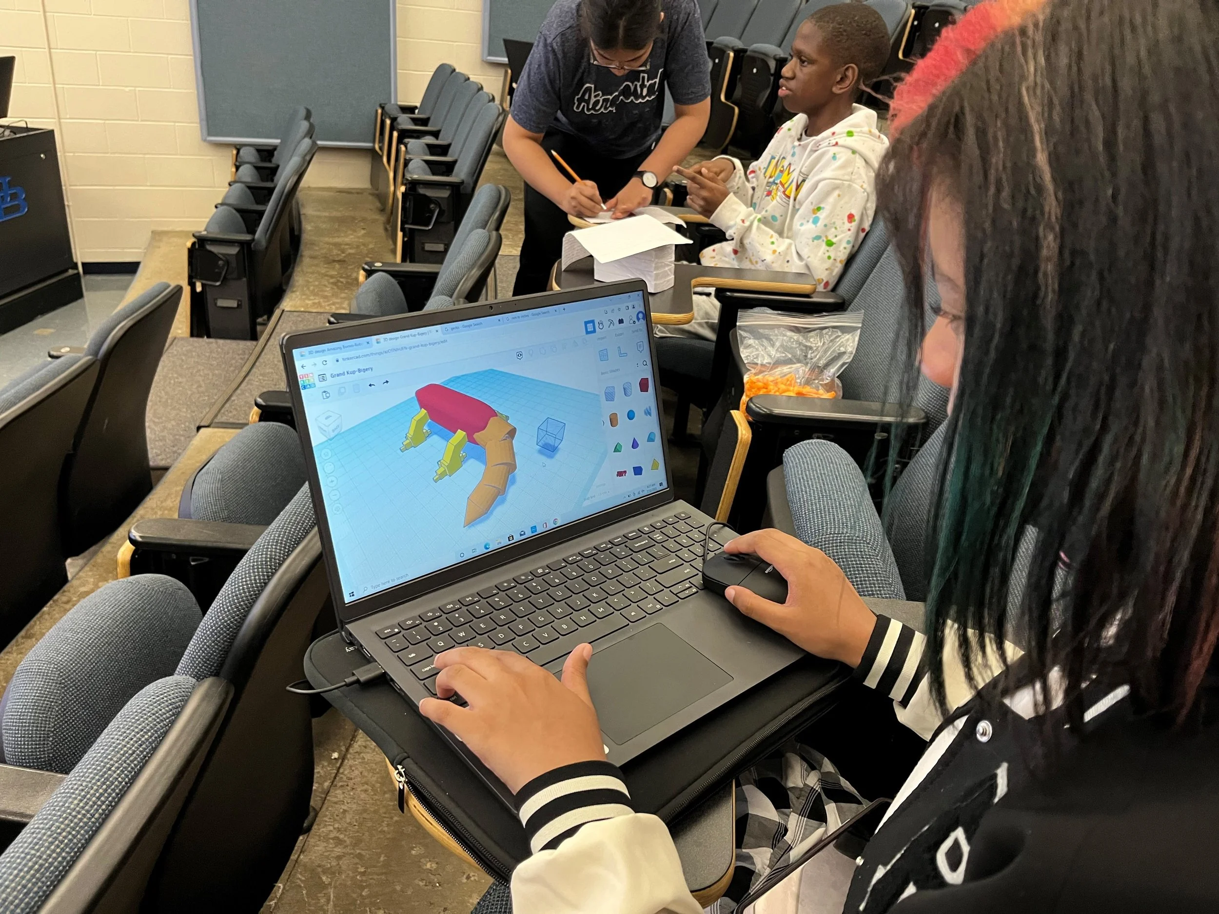 A woman using a laptop with a 3D design of a robot on the screen, in a classroom with other students and empty seats.