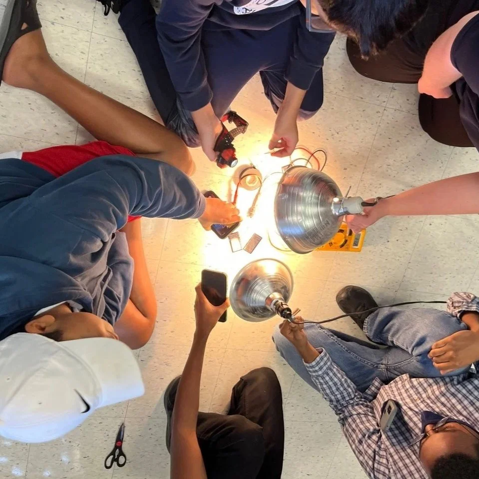 Group of students gathered around a science experiment setup on the floor, with electrical equipment, wires, and metal lamps, working together on a project.