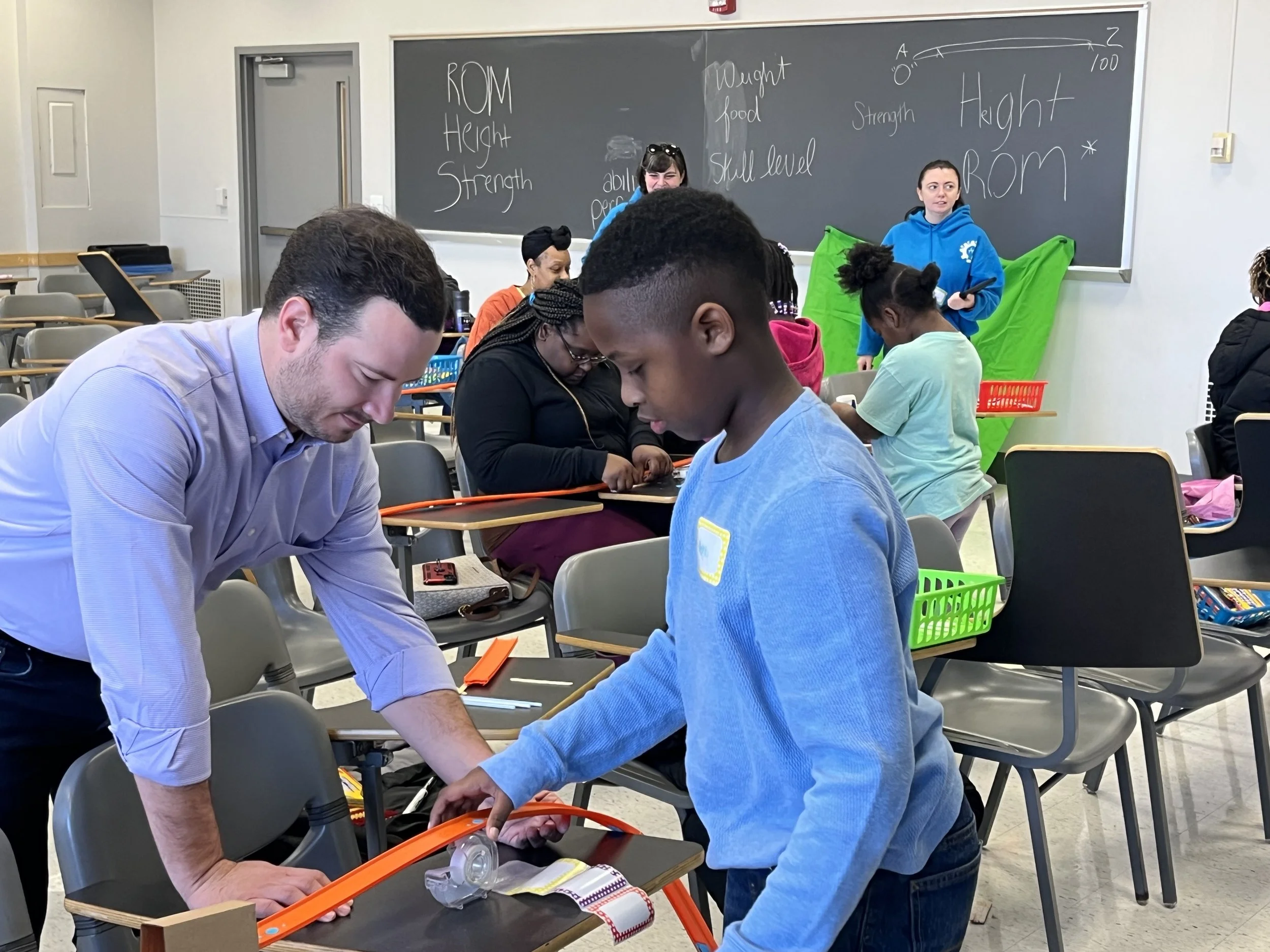 A classroom with children and an adult working on a hands-on project. The blackboard in the background has words and diagrams related to strength measurement and height. Students are engaged with materials on desks.