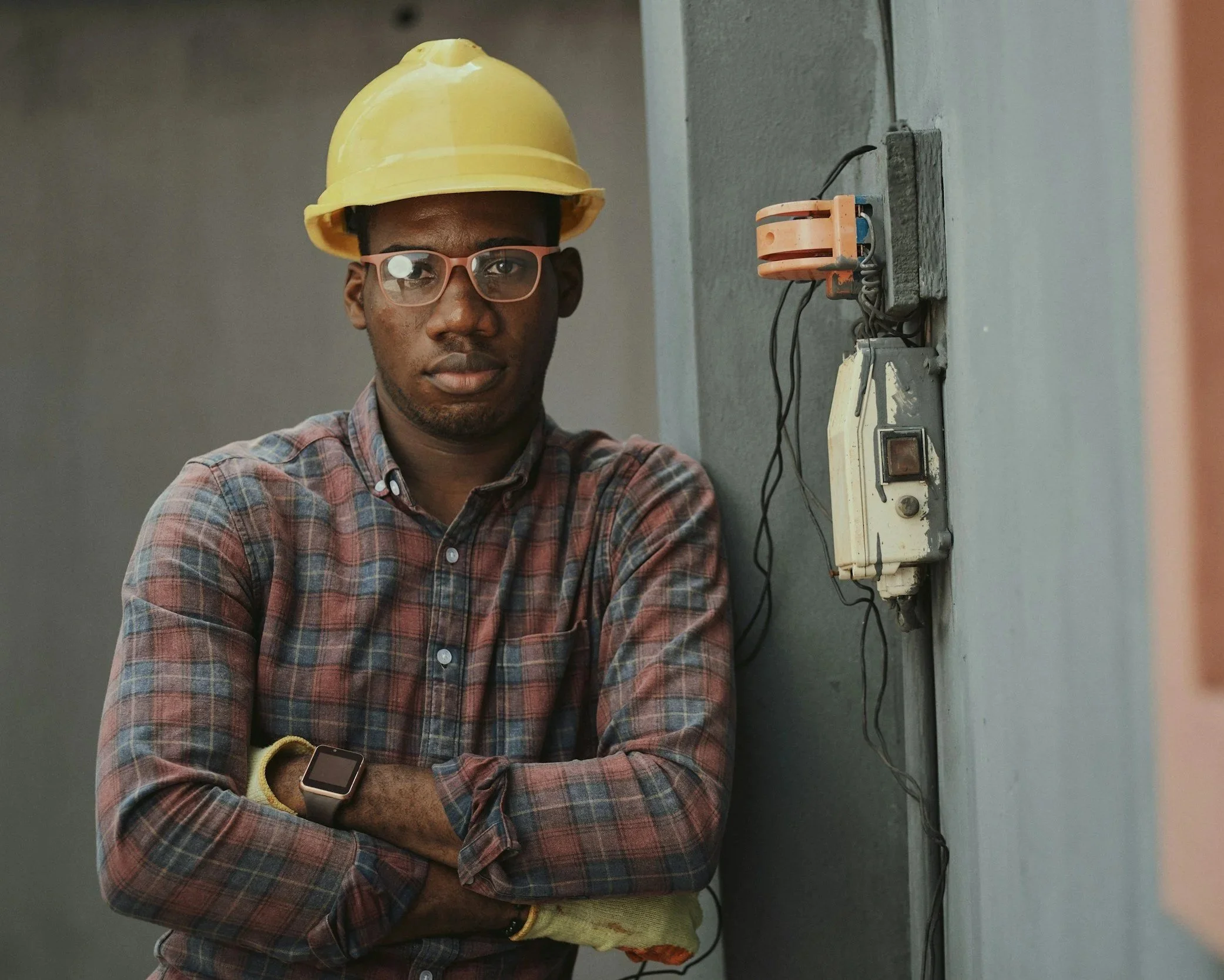 A young man wearing a yellow safety helmet, glasses, and a plaid shirt standing with arms crossed in front of electrical wiring on a gray wall.