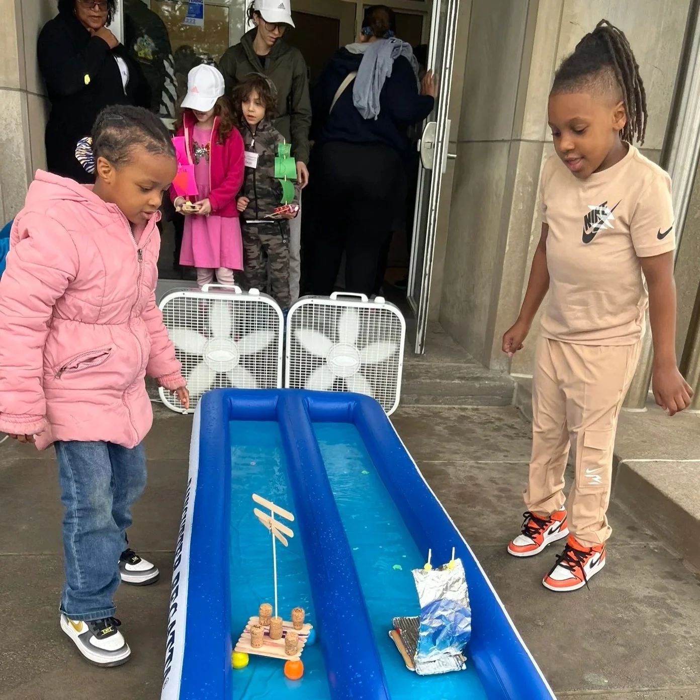 Children playing a tabletop ice hockey game outside a building, with onlookers watching.