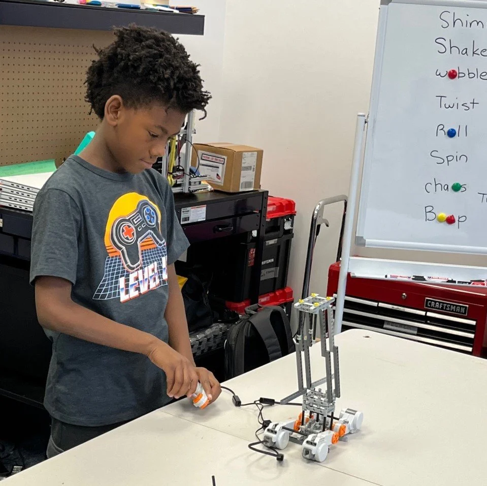 A boy is working on a small robot made of metal and plastic parts on a table in a workshop or classroom setting.
