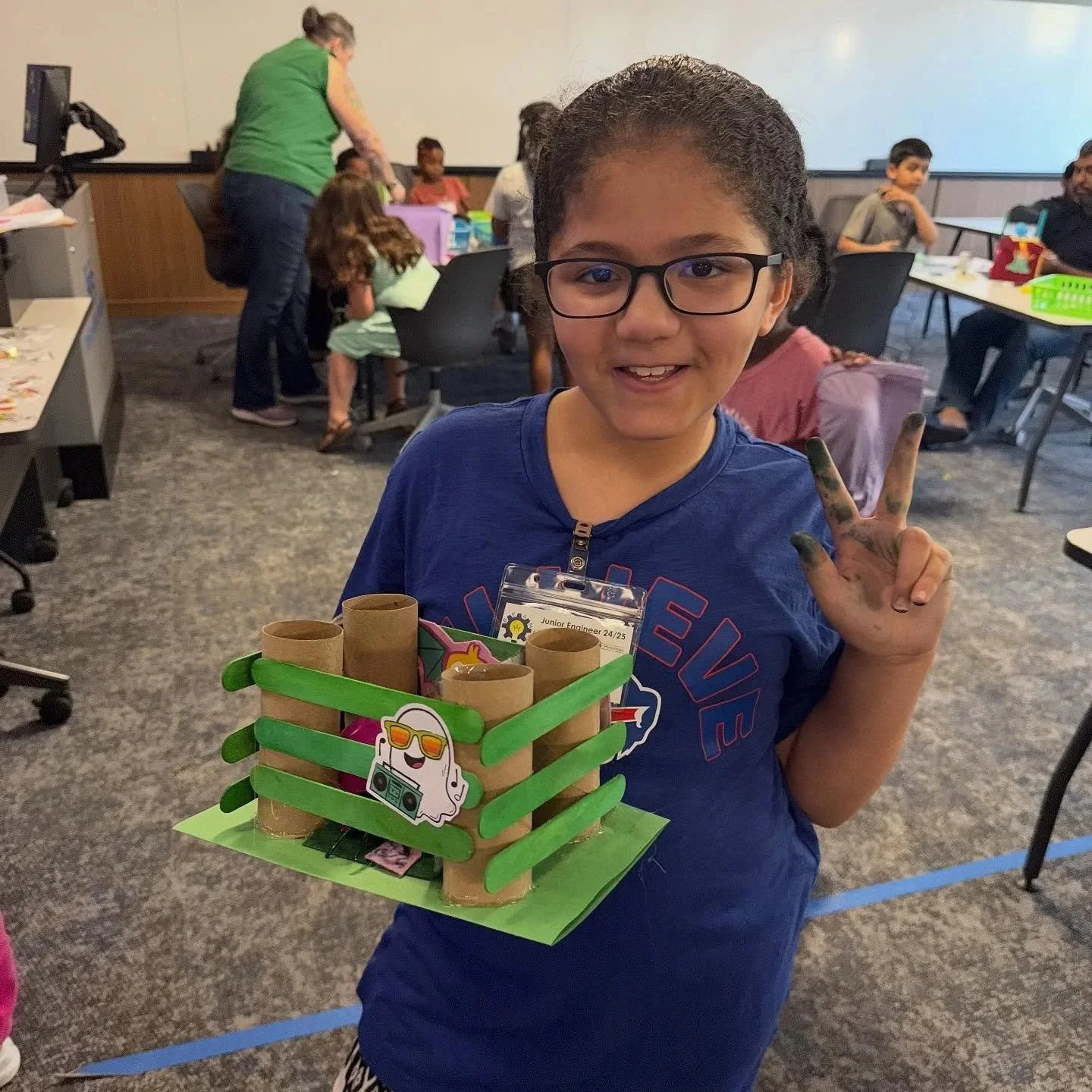 A young girl wearing glasses and a blue shirt smiling and holding a homemade craft project with cardboard tubes and colorful decorations. She is making a peace sign with her hand covered in black marker and is in a classroom or workshop setting with other children and adults in the background.