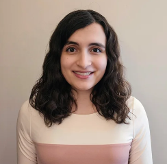 A woman with long curly dark hair smiling at the camera, wearing a white and light pink top, standing against a beige background.