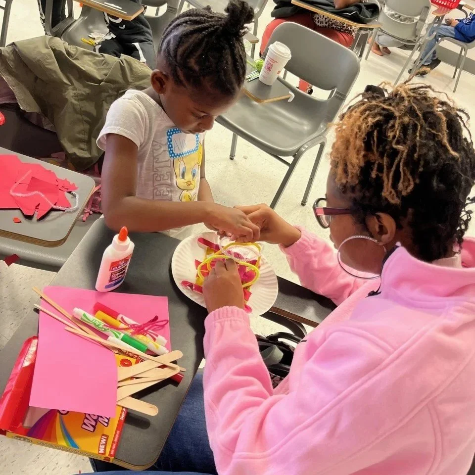 Two children are sitting at a table in a classroom, crafting with colorful ribbons and glue. The girl on the left has braided hair and is wearing a T-shirt with Tweety Bird, while the girl on the right has curly, blonde-tipped hair, glasses, and is dressed in a pink hoodie. Craft supplies and paper are on the table.