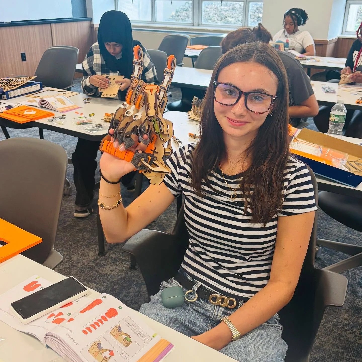 Young woman with glasses smiling and holding a robotic hand model in a classroom setting, with other students working in the background.