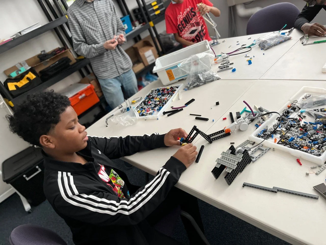 A young boy is assembling a LEGO mechanical structure at a table in a workshop or classroom, with various LEGO pieces and tools spread out on the table. In the background, two other kids are standing and working, surrounded by shelves with boxes and supplies.