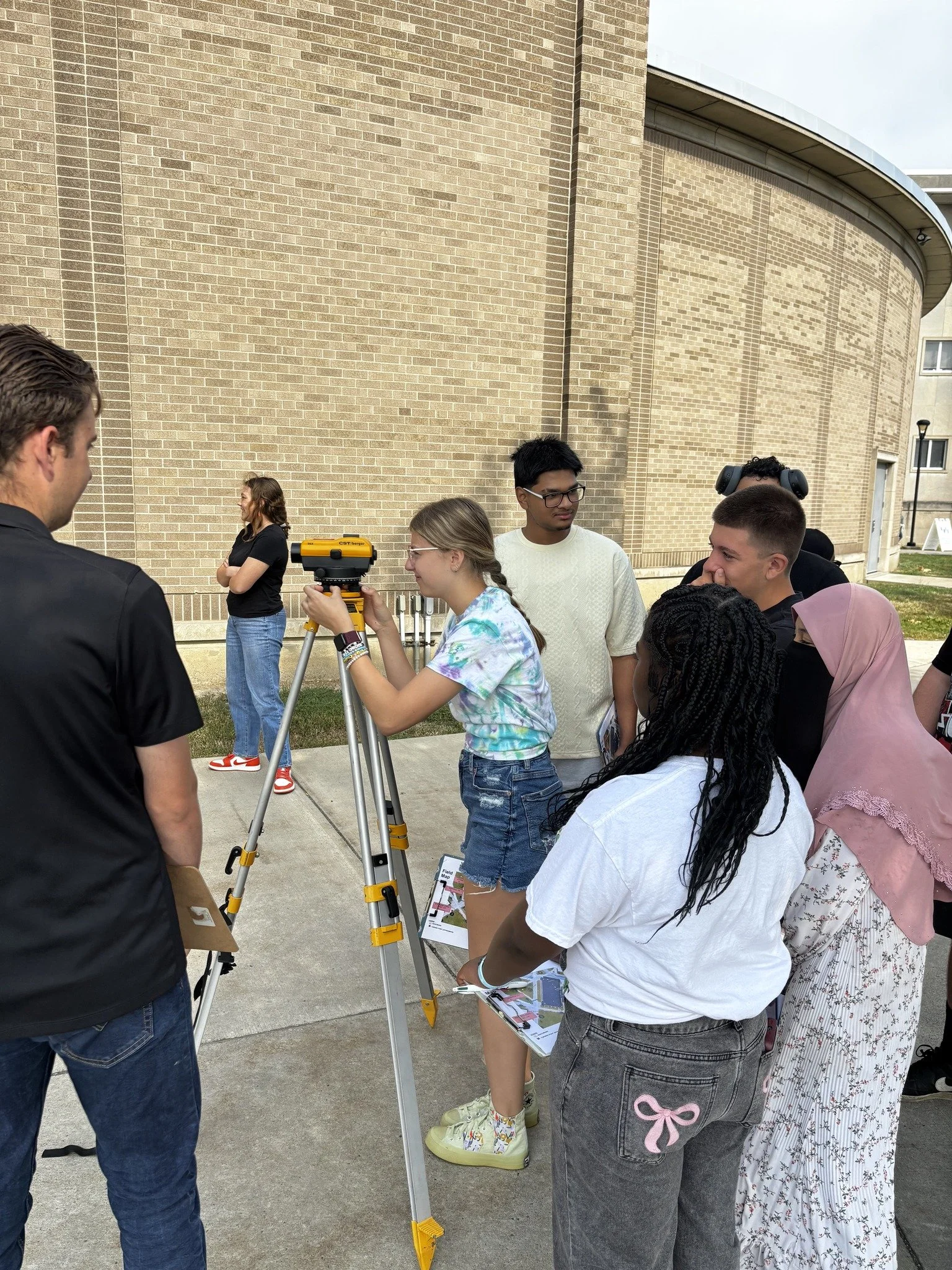 A group of young people gathered around a girl operating a surveying instrument outside a brick building, with some people in the background observing.