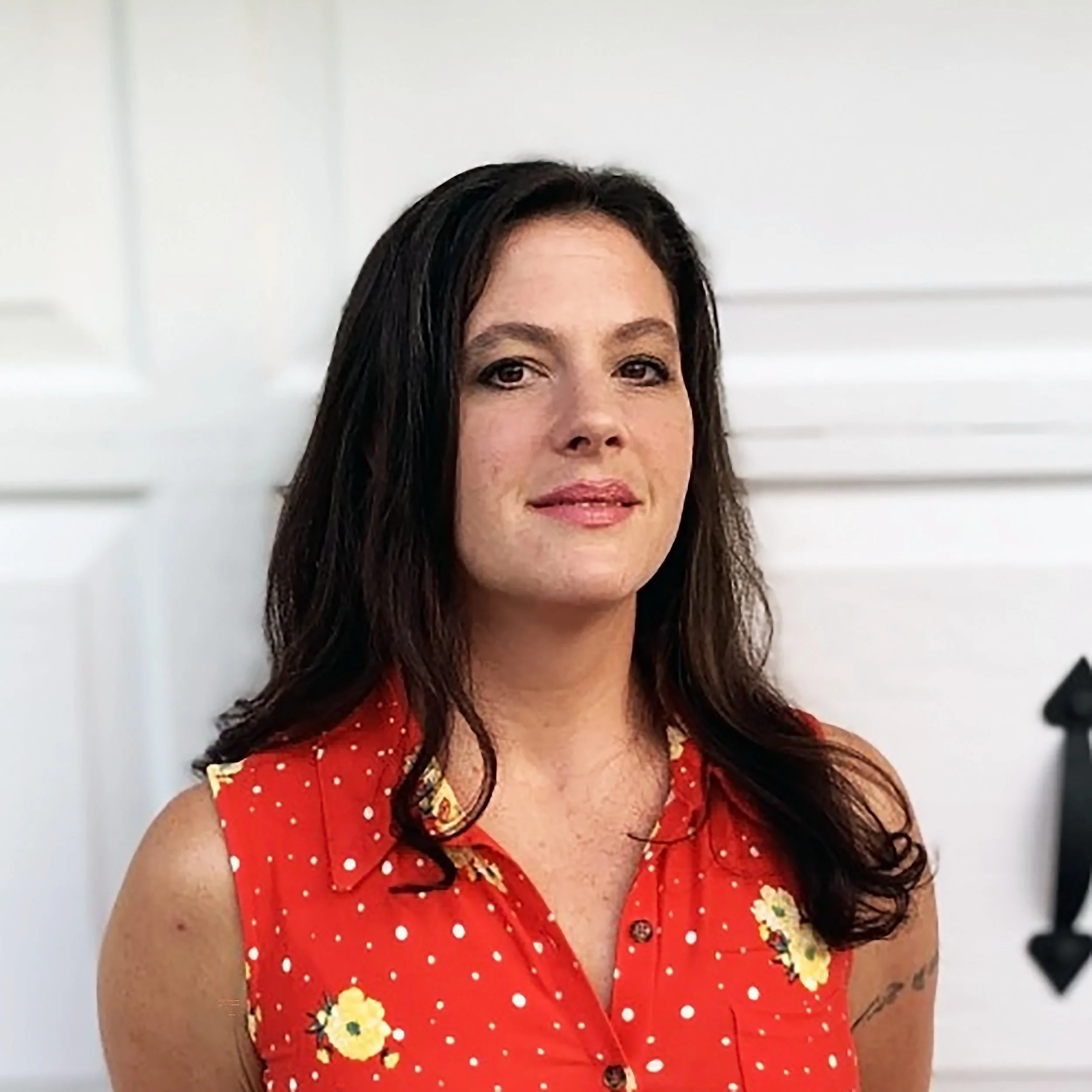 Portrait of a woman with dark wavy hair wearing a sleeveless red dress with yellow floral patterns and white polka dots, standing in front of a white door.