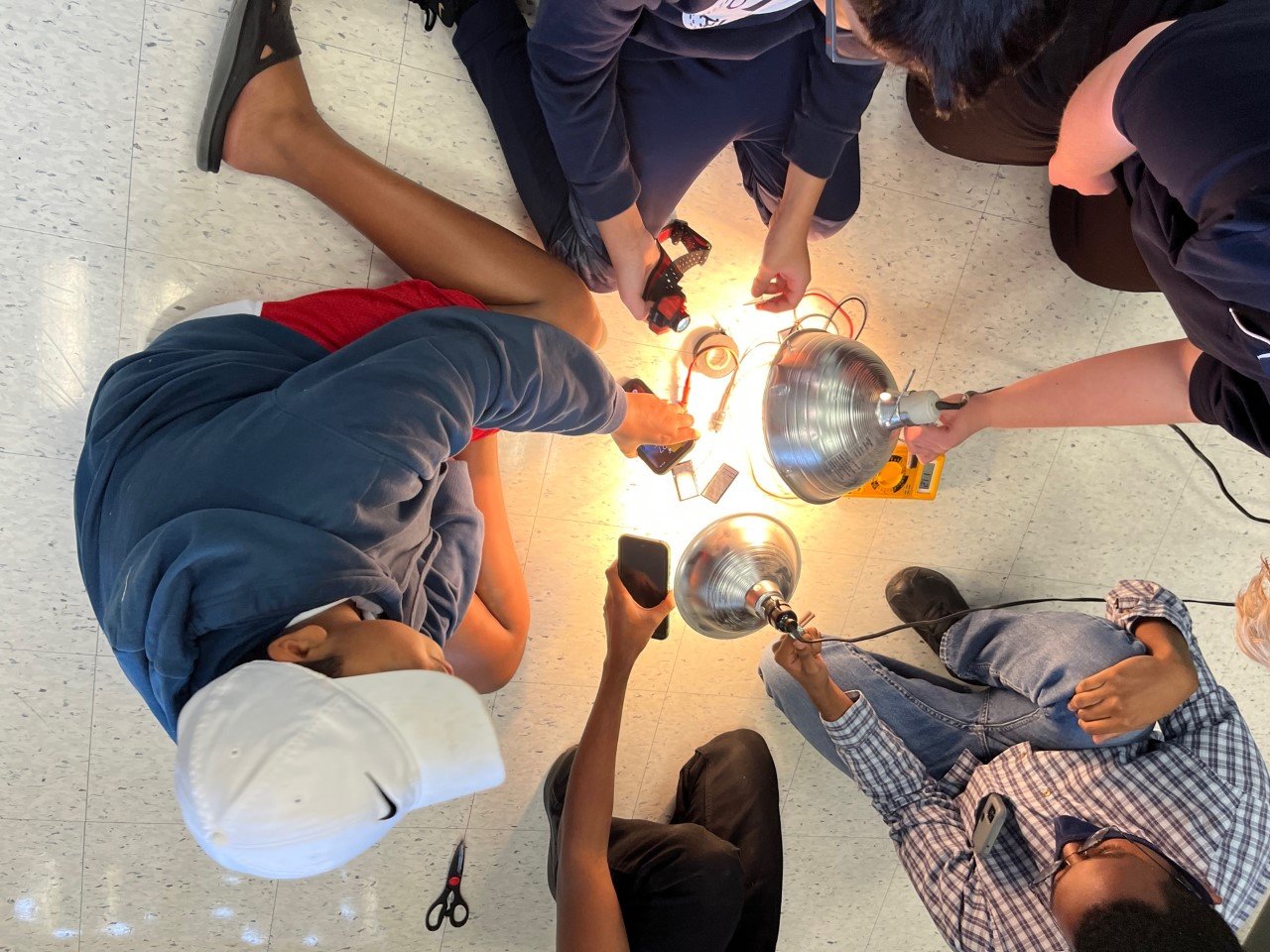A group of children working together on a science project involving electrical components and light bulbs, seen from above.