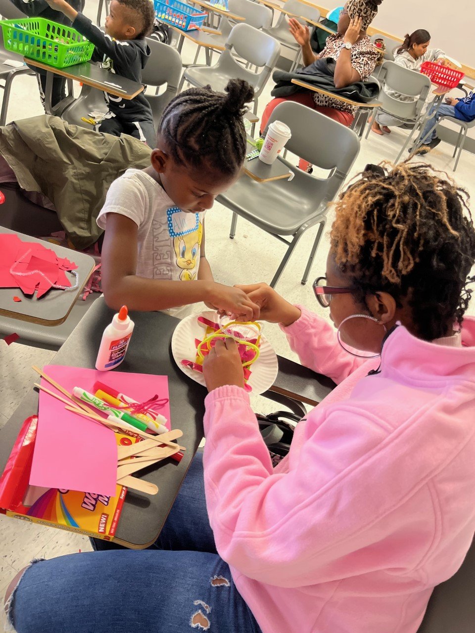 Two girls are sitting at a desk in a classroom, working together on a craft project with colorful materials. Other children are visible working and playing in the background.
