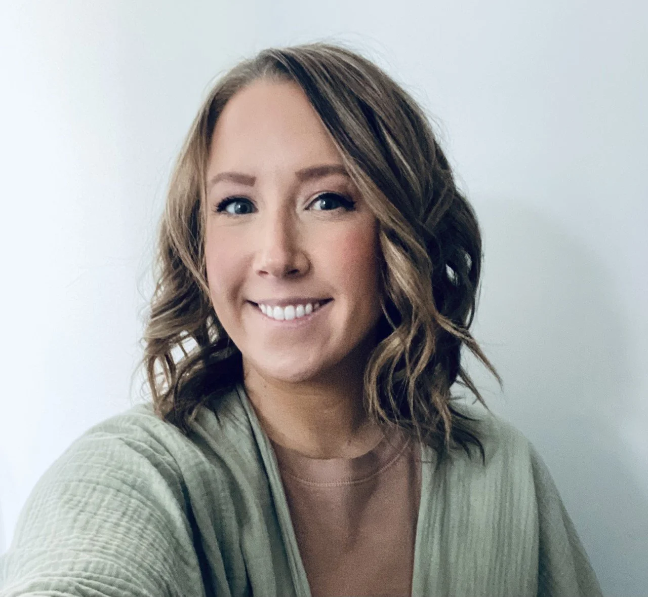 A woman with light brown, shoulder-length, curly hair smiling at the camera against a plain light-colored background.
