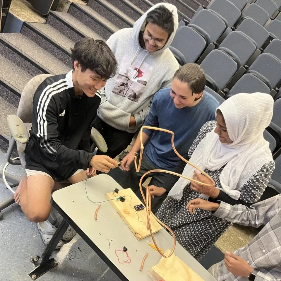 A group of five young people gathered around a table, engaged with a DIY electronics project involving wires and small electronic components.