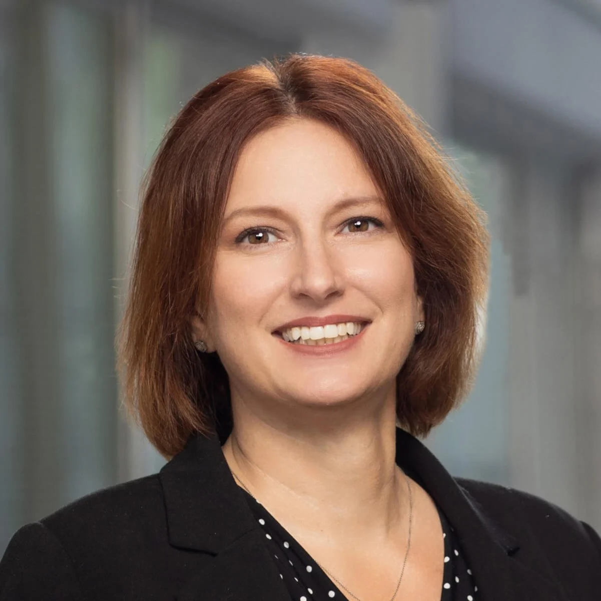 Professional woman with shoulder-length reddish hair smiling, wearing a black blazer and polka dot top, in an indoor office or corporate setting.
