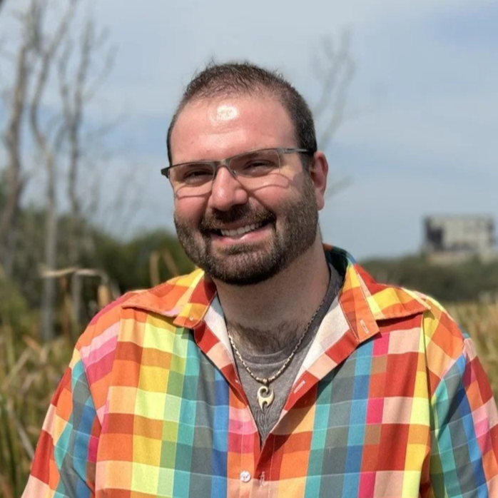 A smiling man wearing glasses and a colorful plaid shirt outdoors in front of a blue sky and leafless trees.