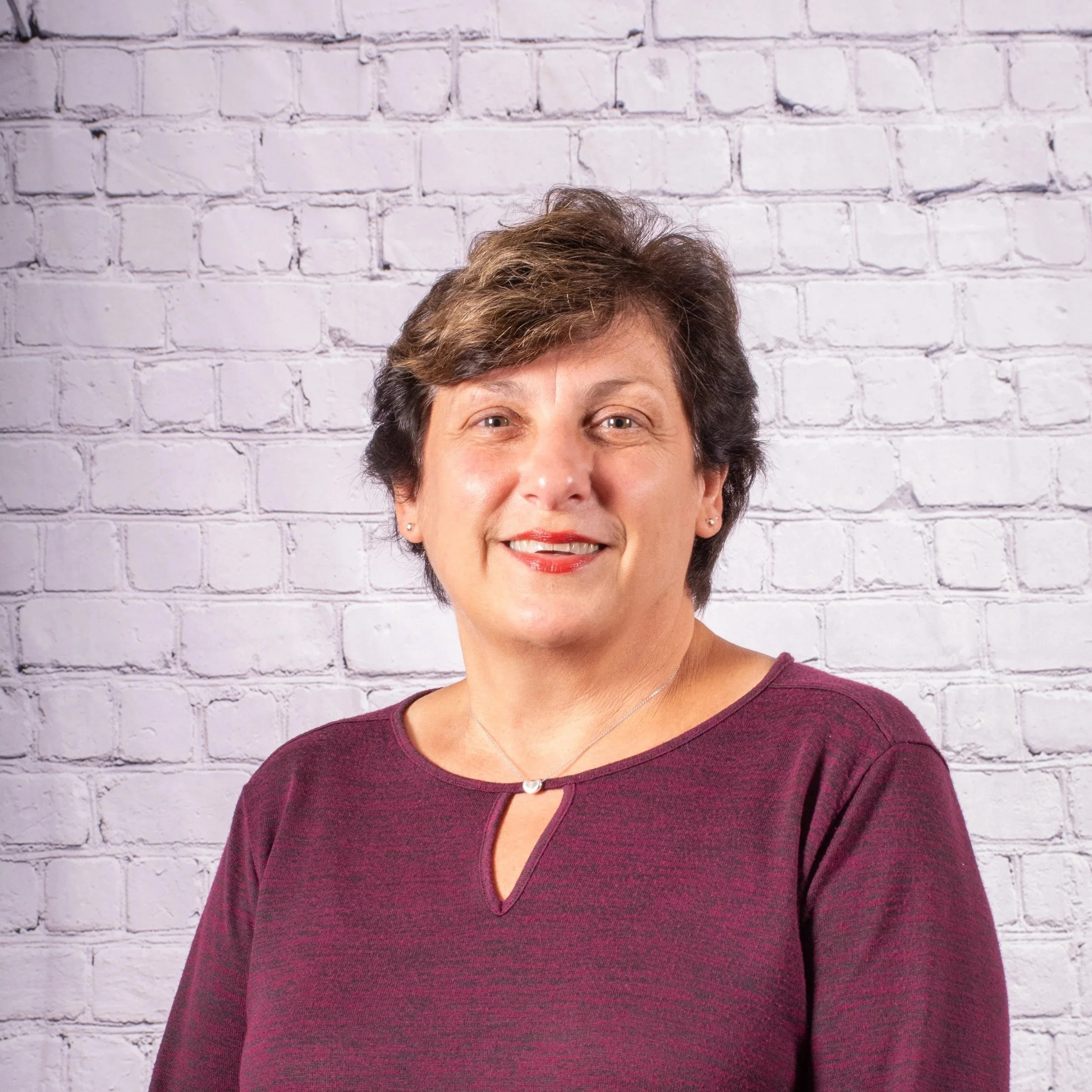 A woman with short brown hair and a maroon top standing in front of a white brick wall.