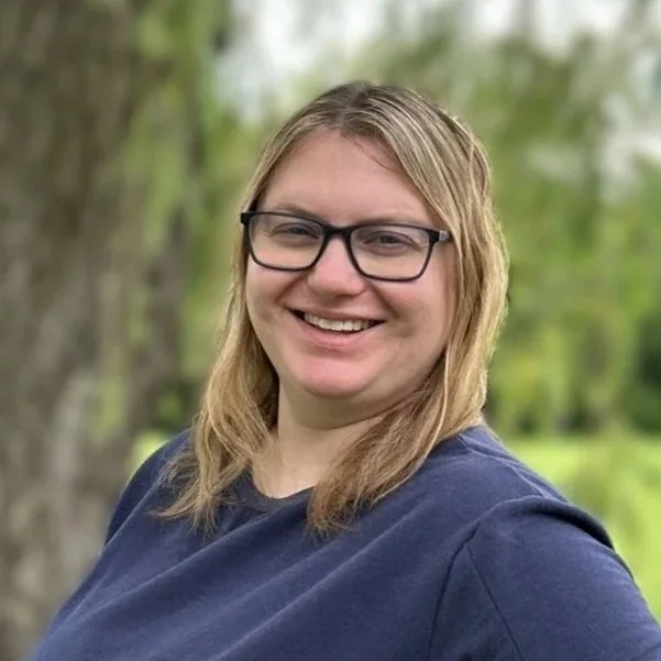 A woman with blonde hair wearing glasses and a navy blue shirt, smiling outdoors with trees and greenery in the background.