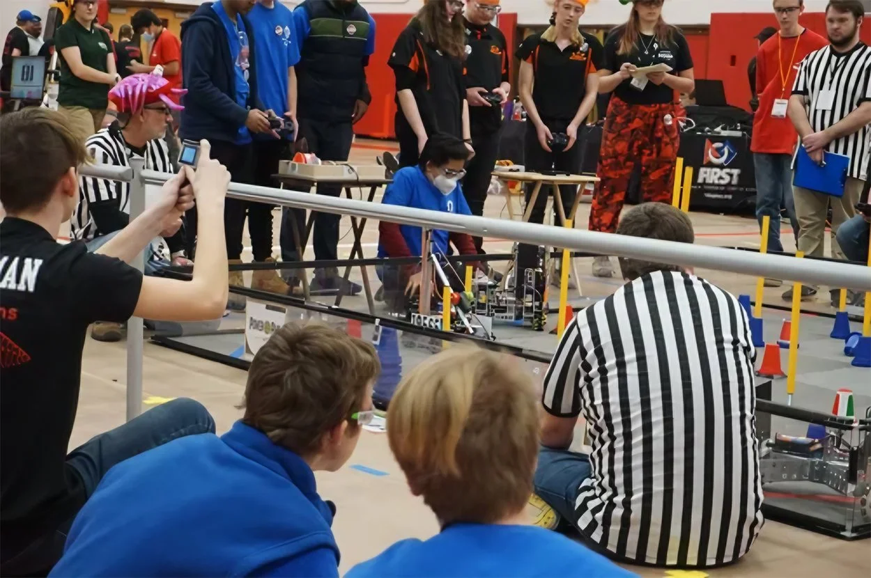 Students and judges participating in a robotics competition in a gymnasium, with robots, barriers, and cones on the floor.