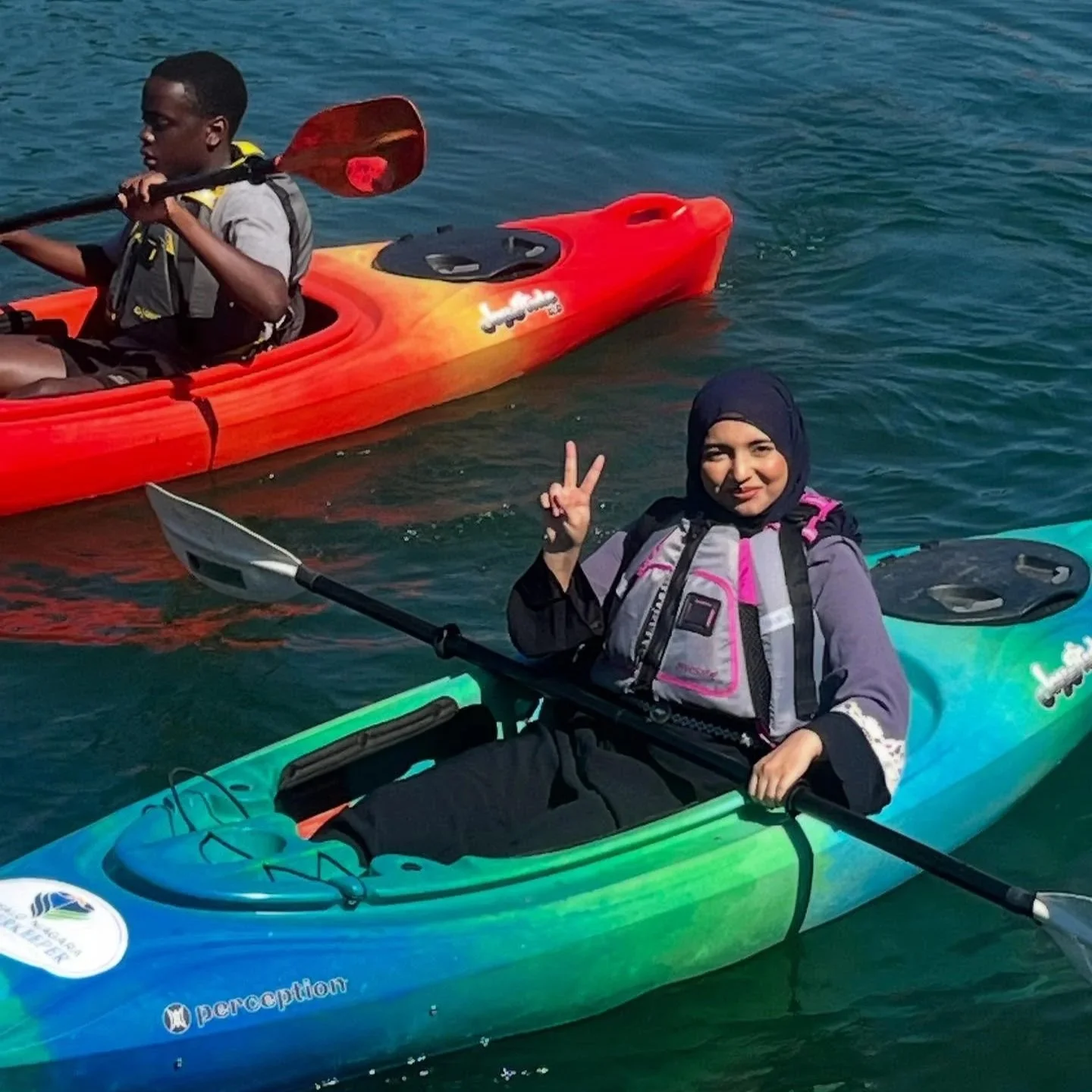 Two people in kayaks on the water, one woman smiling and making a peace sign, the other person paddling in a red kayak.
