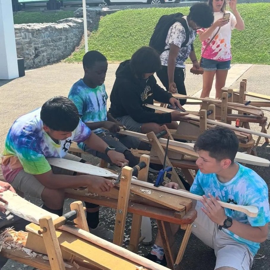 Group of young people operating woodworking lathes outdoors, learning woodcraft with a woman supervising.