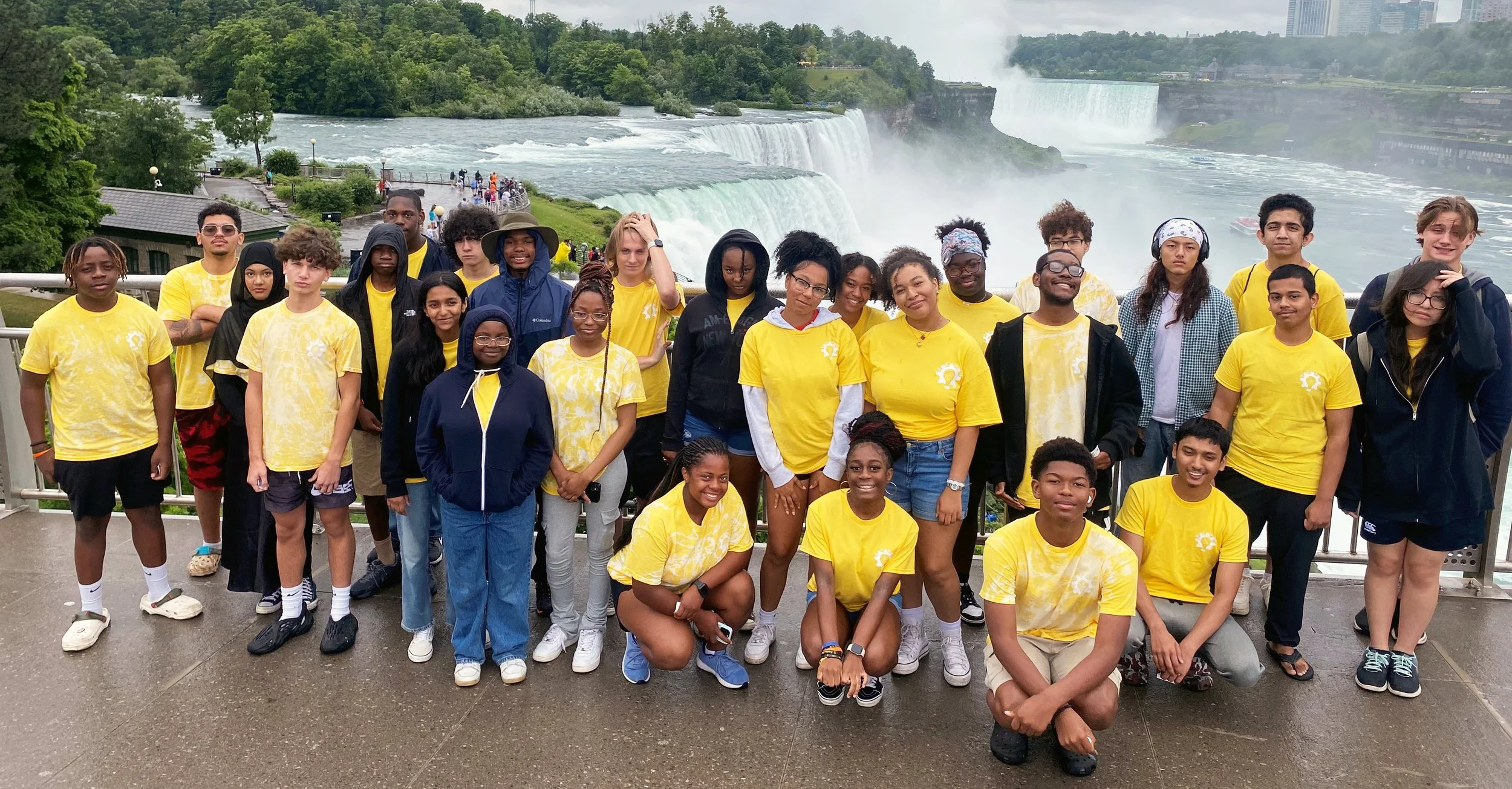 A group of diverse young people wearing yellow shirts stands in front of Niagara Falls during a rainy day.