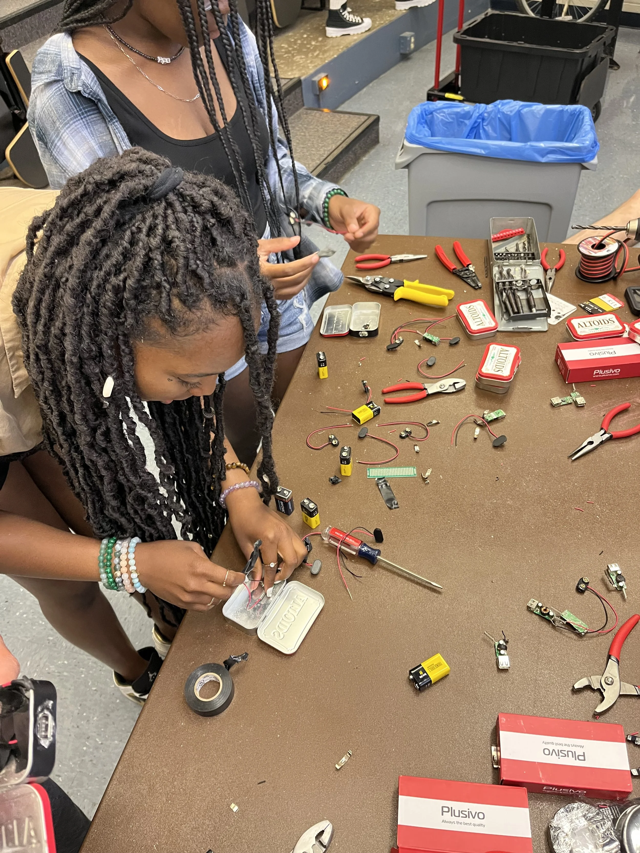 Two young women working together on electronics repair tools on a cluttered brown work table.