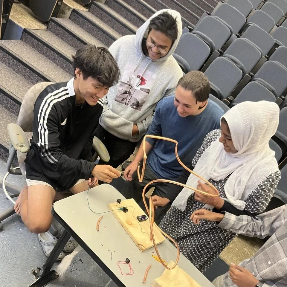 A group of five diverse young people gathered around a small table, engaging in a hands-on science activity with wires, batteries, and bulbs, in a lecture hall or auditorium with empty seats.