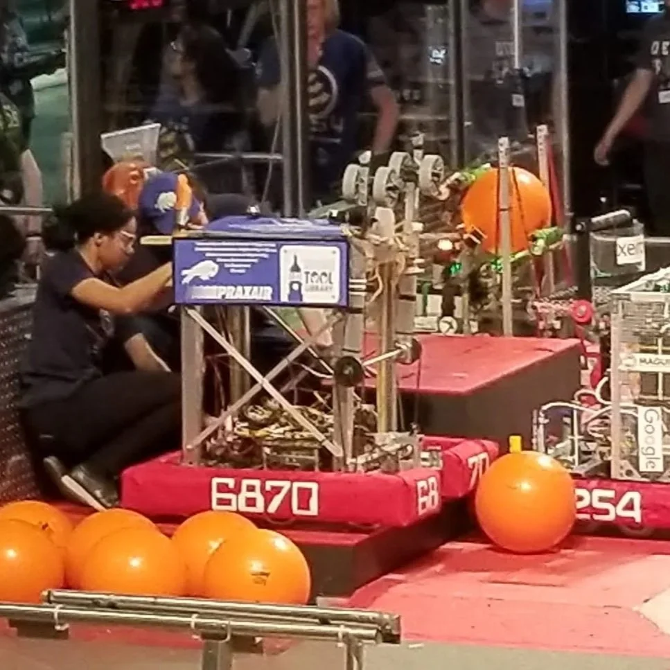A person with glasses and dark hair playing a robot game at a carnival, with large orange balls and various equipment in the background.