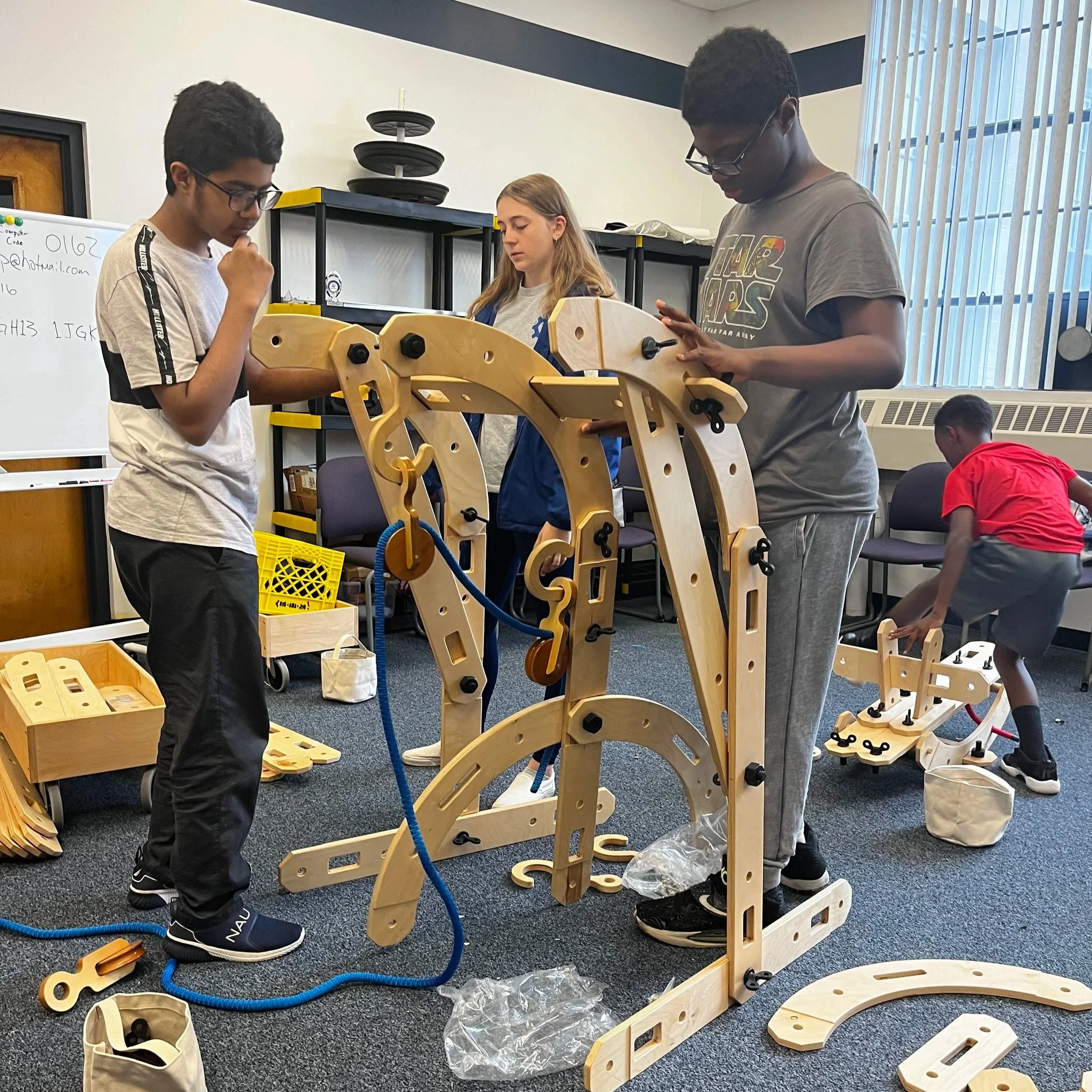 Three children working on assembling a wooden mechanical structure in a classroom setting, with another child in the background playing with a smaller wooden contraption.