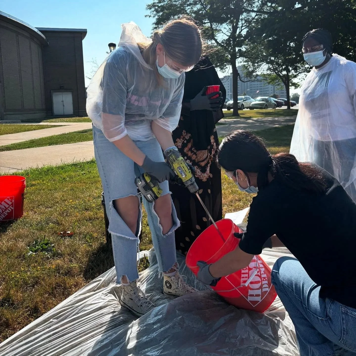 Group of four people, all wearing masks and gloves, working together outdoors with tools and a red bucket on a plastic sheet on the grass. One woman is using a power drill, others are assisting.