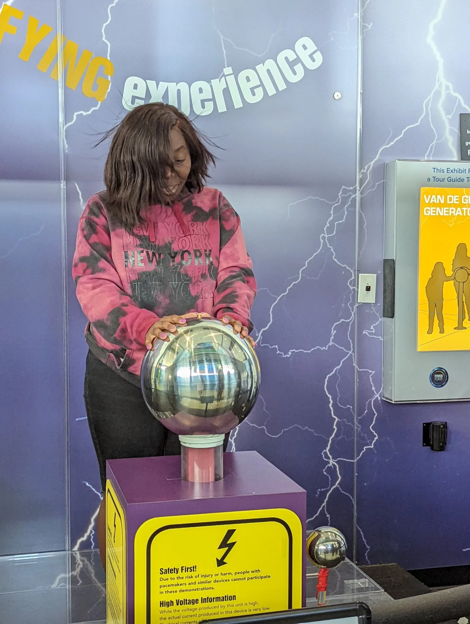 A young girl is touching a large, shiny, metal sphere that is part of a science exhibit on high voltage electricity, surrounded by lightning-themed decor.