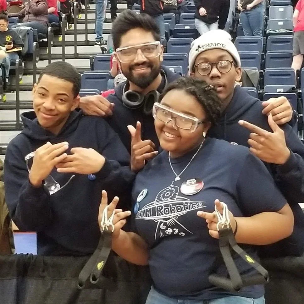 Four people standing together at a robotics competition, wearing glasses and team shirts, with a crowd in the background.