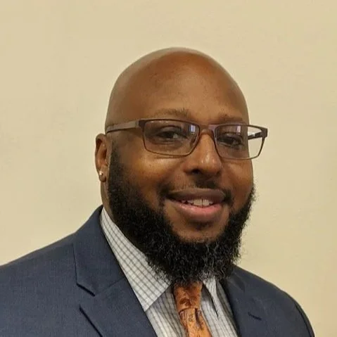 Headshot of a smiling man with a beard, wearing glasses, a blue suit, and an orange tie, against a plain light-colored background.