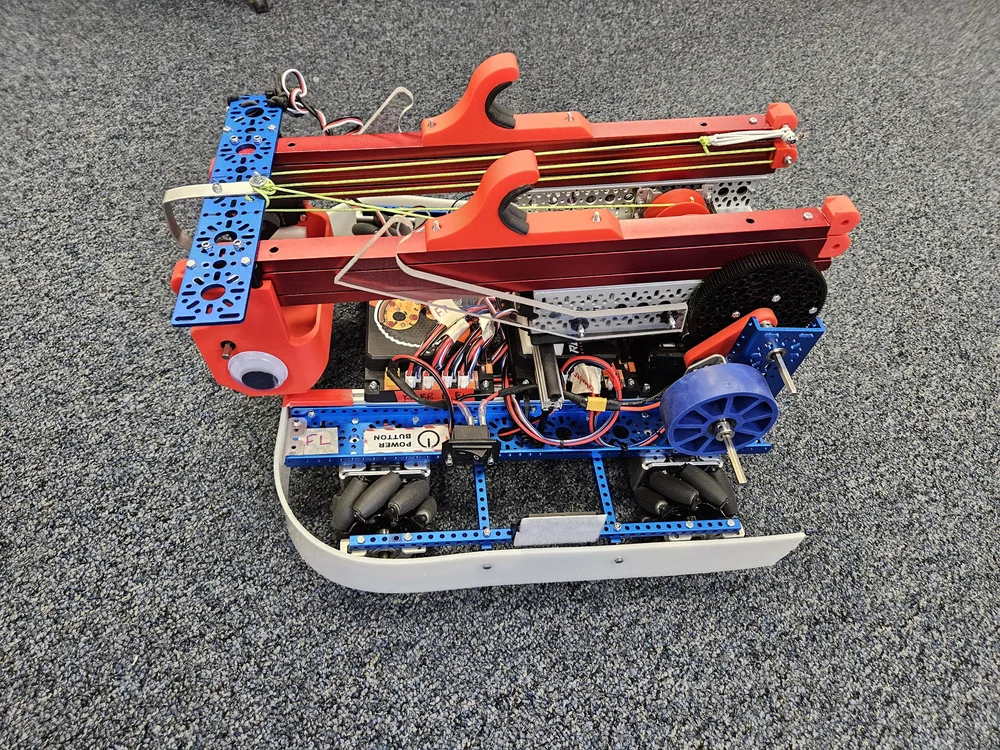 A partially assembled robot with red, blue, and gray metal parts, wheels, and wiring, placed on a gray carpeted surface.