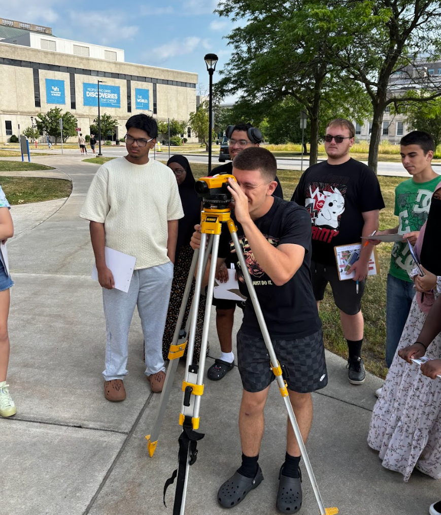 A group of young people gathered outdoors around a man using a theodolite or surveyor instrument on a tripod, with some holding notebooks and papers. Trees and a large building with banners are in the background.