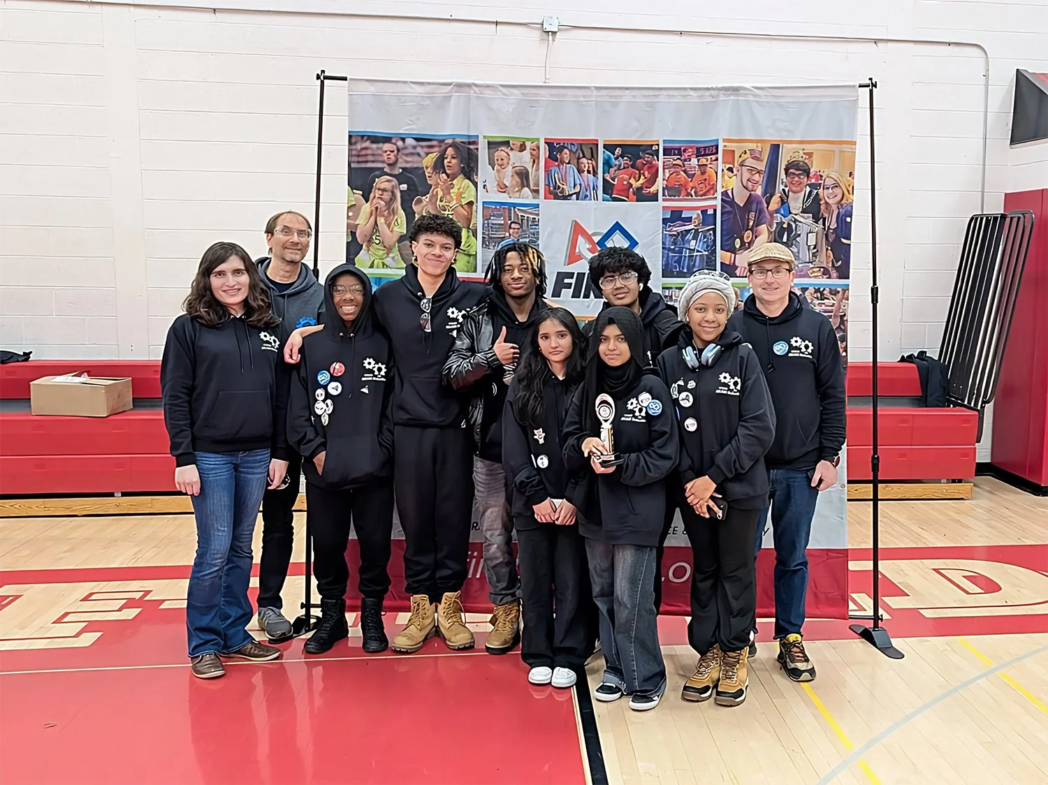 A group of nine people, some wearing black hoodies with logos, stand together on a gymnasium floor in front of a backdrop with photos of people and a logo. One person in the front holds an award.