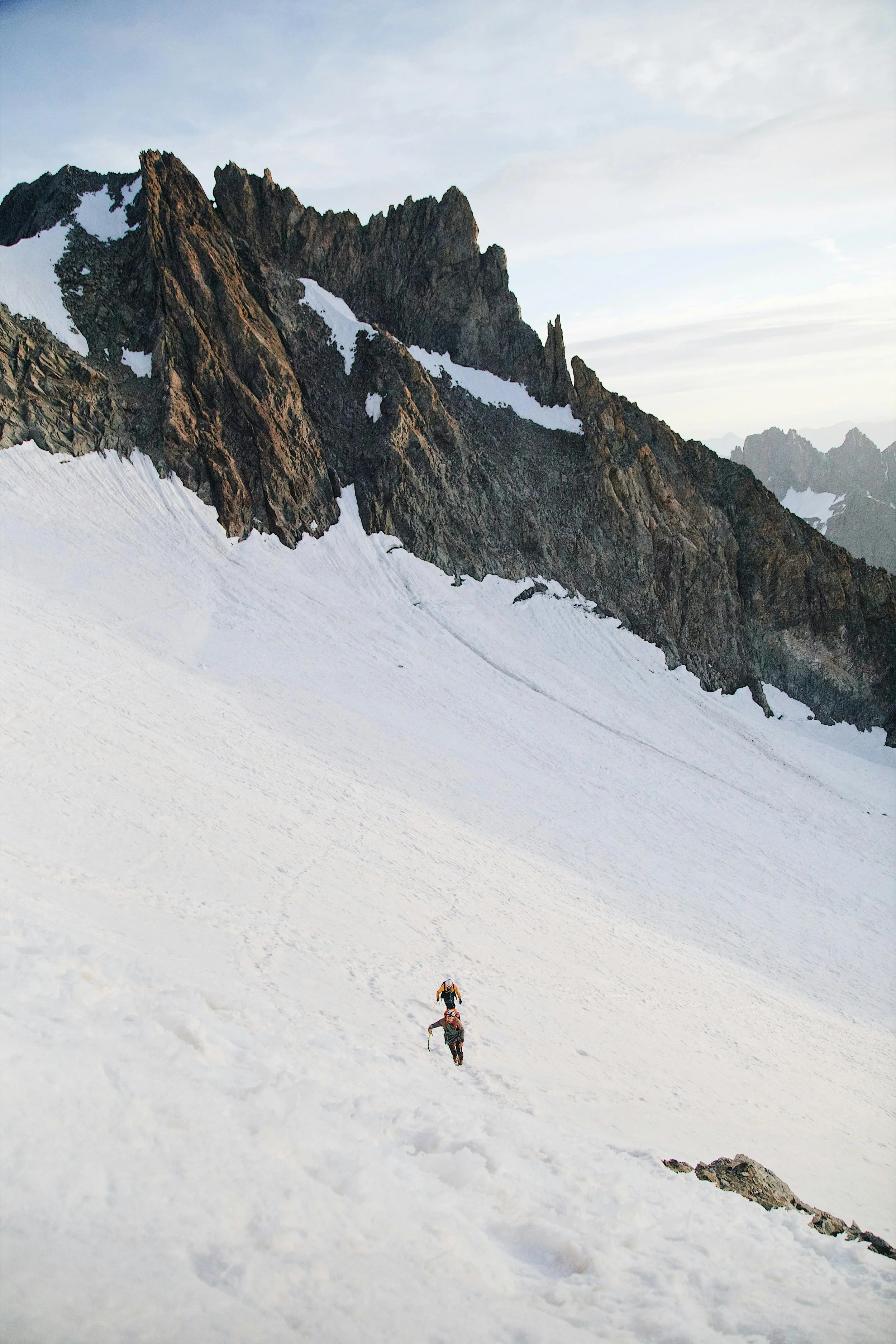 Two climbers ascending a snow-covered mountain slope with rocky peaks in the distance under a cloudy sky.