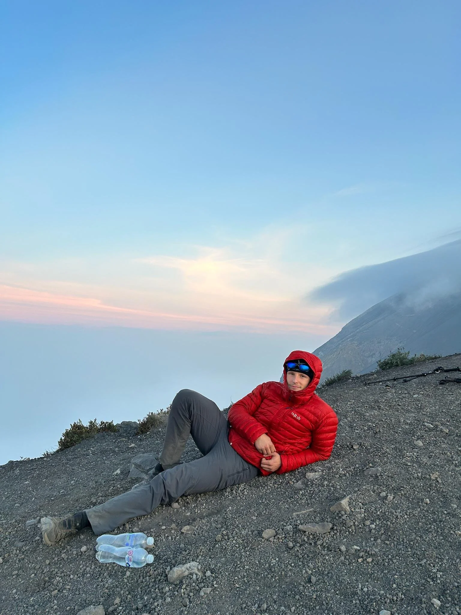 A person wearing a red jacket, black pants, and sunglasses lounging on a rocky mountain slope near the edge, with a scenic background of mountains, sky, and clouds.