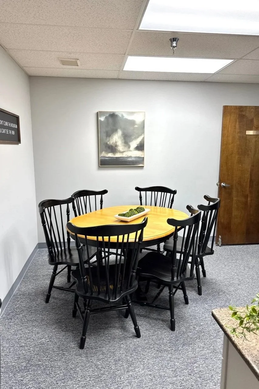 A small conference room with a round wooden table surrounded by six black chairs. The table has a decorative dish with green ornamental balls. A gray and white abstract painting hangs on the white wall at the back, and a closed wooden door is on the 