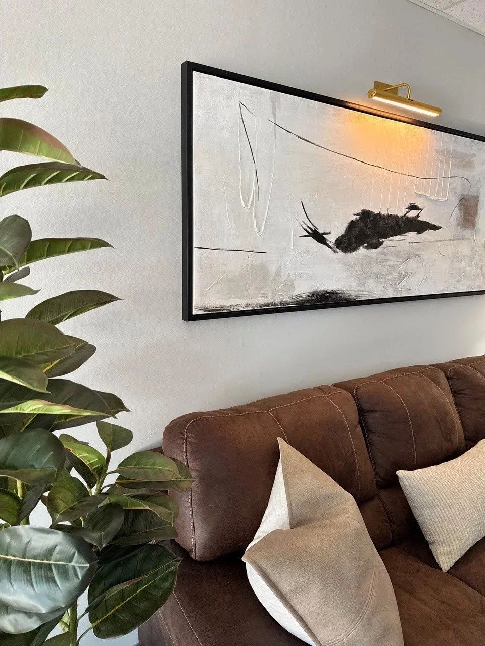 Living room with a brown leather sofa, beige pillows, a large abstract black and white painting on the wall, a brass picture light, and a tall green plant.
