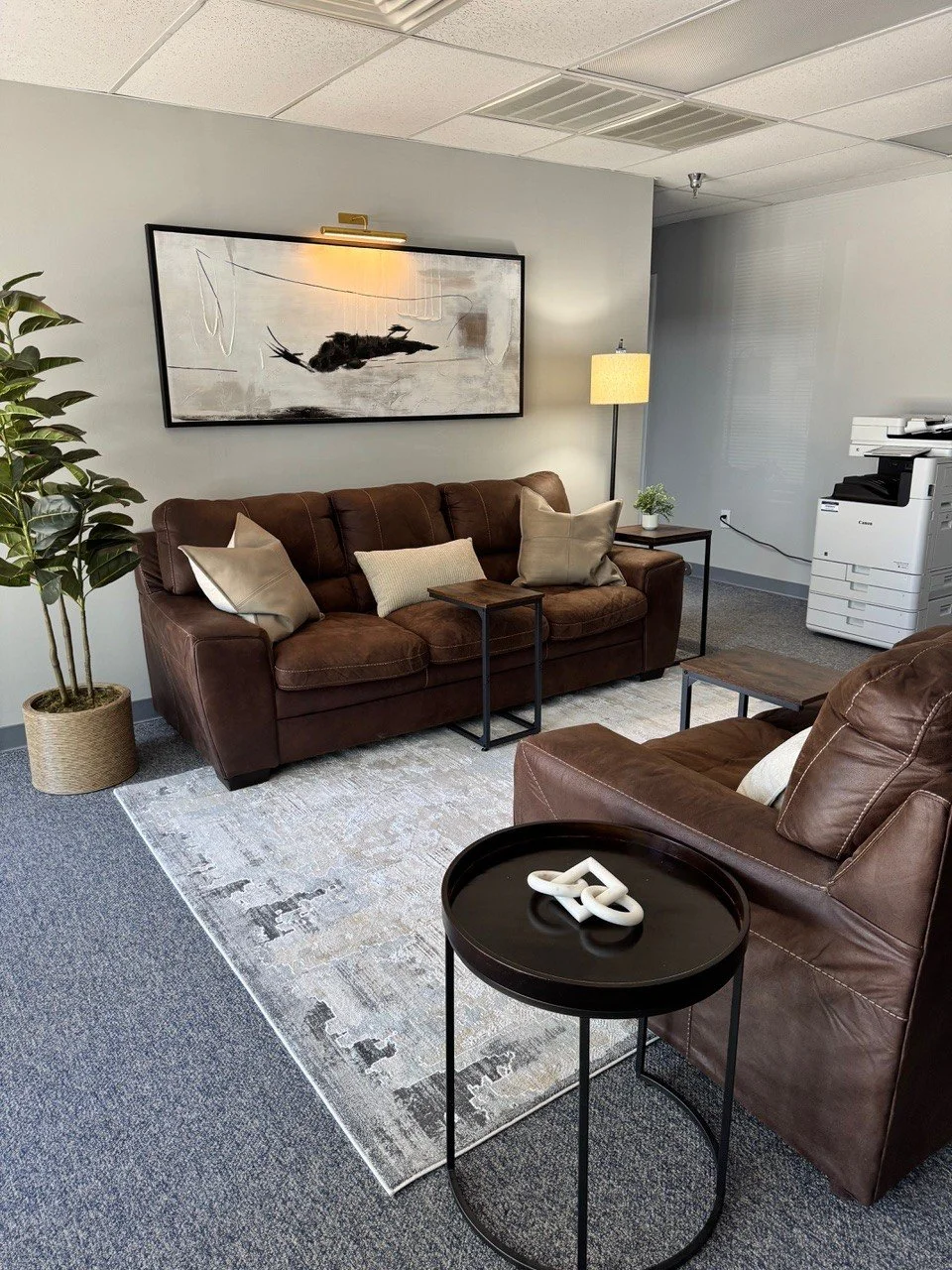 Office waiting area with two brown leather sofas, a black side table with a white sculpture, a patterned area rug, a tall potted plant, a floor lamp, wall art, and a copier machine.