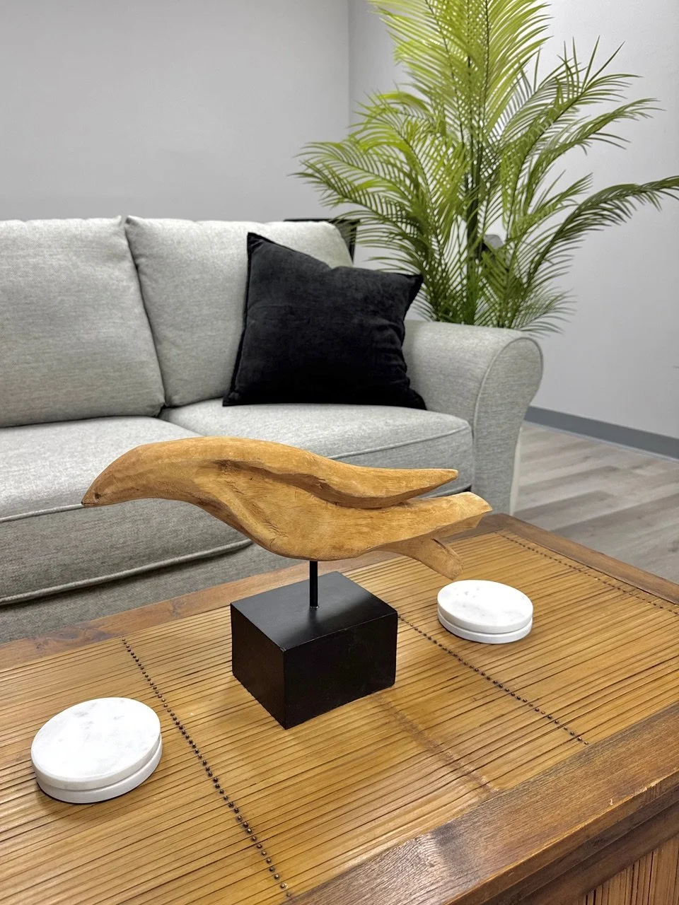 A wooden decorative object resembling a bird on a black stand placed on a wooden table, with a gray sofa, black pillow, and a green potted plant in the background.
