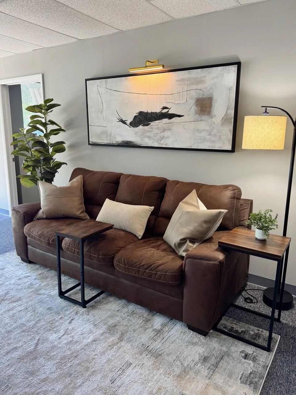 Living room with a brown leather sofa, beige pillows, a small wooden side table, a floor lamp, a large abstract painting on the wall, a potted plant, and a window with blinds.