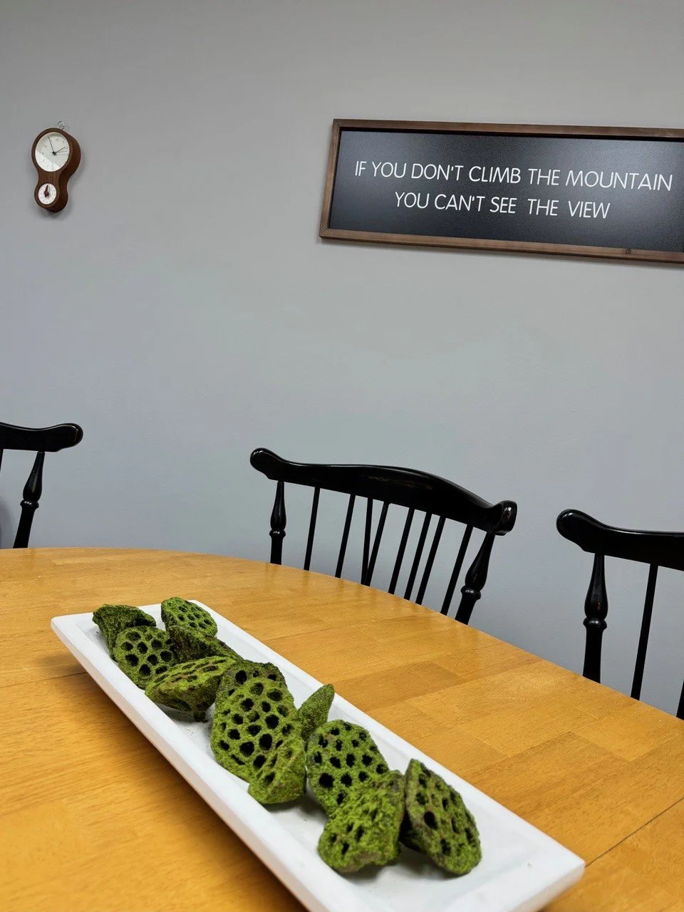 A white rectangular platter with pieces of fried okra on a wooden table in a room with black chairs, a wall clock, and a sign that reads 'If you don't climb the mountain you can't see the view'.