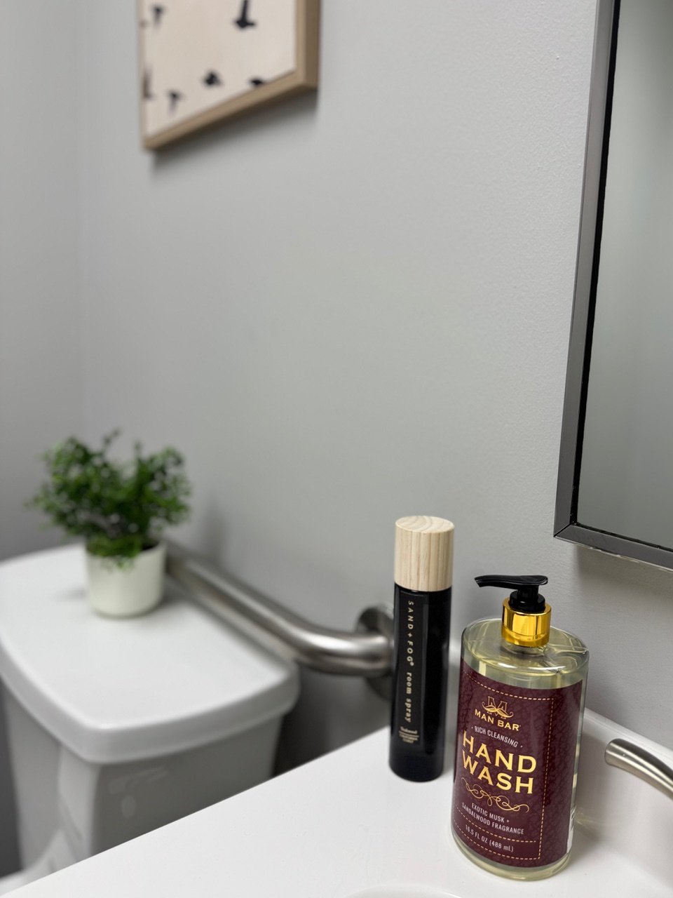 A white bathroom sink with a bottle of hand wash, a small container of sand fog perfume, and a small potted green plant on top. A mirror is partially visible on the right side. The background includes a gray wall, a soap holder, and a framed art piec